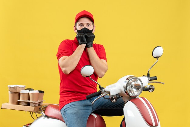 Top view of scared young adult wearing red blouse and hat gloves in medical mask delivering order sitting on scooter on yellow background