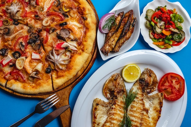 Вид сверху морепродукты микс пицца с осьминогом грибы крабовое мясо томатный сыр жареная рыба с ломтиком лимона красный лук и овощной салат на столе