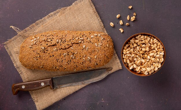 Top view of seeded baguette and knife on sackcloth with bowl of corns on maroon background with copy space