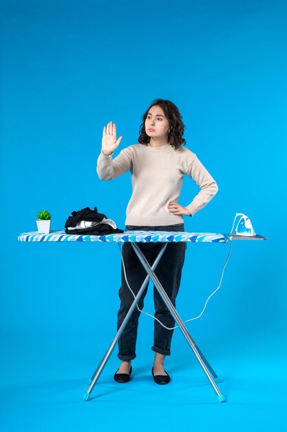 Top view of serious young girl standing behind the ironing board making stop gesture on blue wave background