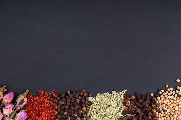 Top view of a set of spices and herbs tea rose buds red chili pepper flakes black peppercorns anise seeds and clove on black background with copy space