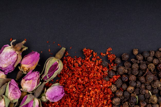 Top view of a set of spices and herbs tea rose buds red chili pepper flakes and black peppercorns on black background
