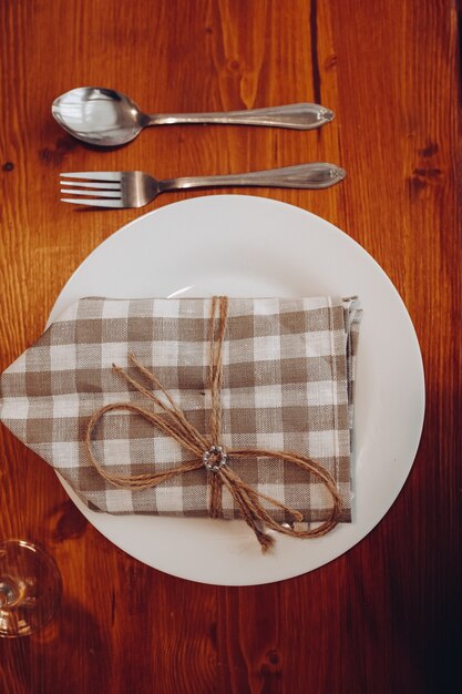 Top view of setting of white ceramic plate with checked brown and white napkin tied with rustic ribbon. Served on brown wooden table.