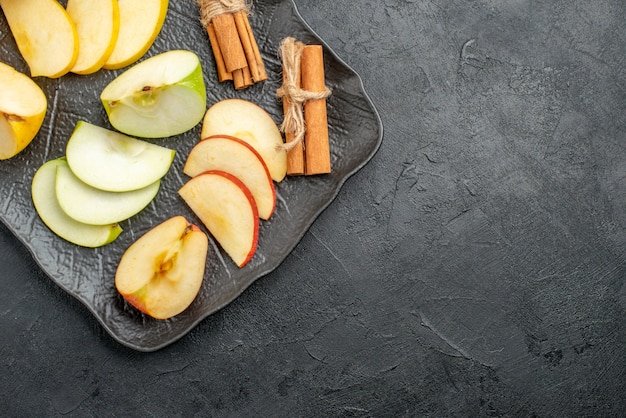 Top view of several types of sliced and whole fresh apples on a black tray and cinnamon limes on the left side on a dark background