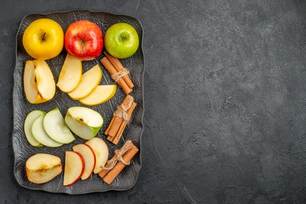 Top view of several types of sliced and whole fresh apples and cinnamon limes on a black tray on the right side on a dark background