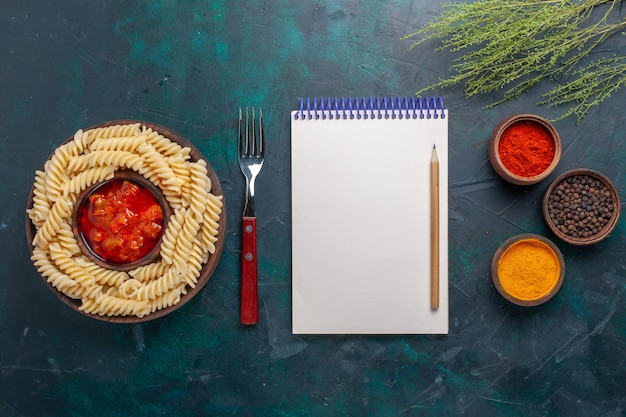 濃い青の背景にメモ帳とさまざまな調味料を使った上面図の形をしたイタリアンパスタ