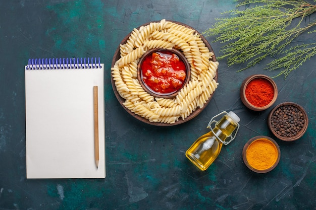 Top view shaped italian pasta with oil notepad and different seasonings on dark blue background