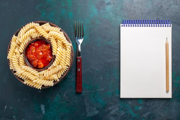 Top view shaped italian pasta with tomato sauce and notepad on dark-blue background