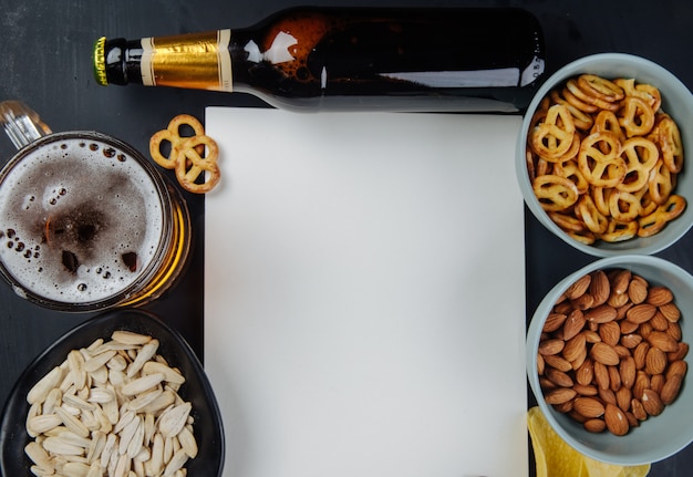 Top view of a sheet of white paper and a bottle of beer with varied beer snacks and a mug of beer on black