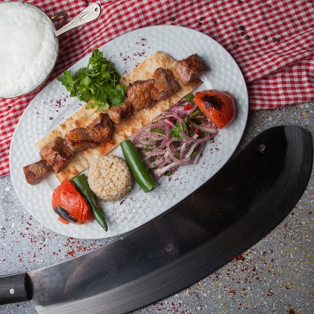 Top view shish kebab with fried vegetables and chopped onion and ayran and knife in white plate