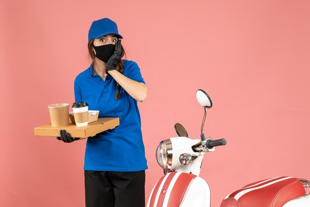 パステル ピーチ色の背景にコーヒーの小さなケーキを保持しているオートバイの隣に立っている医療マスク手袋を着てショックを受けた宅配便の女の子のトップ ビュー