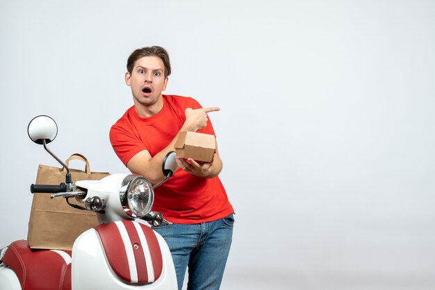 Top view of shocked delivery guy in red uniform standing near scooter giving order pointing back on white background