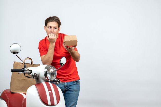 Top view of shocked delivery guy in red uniform standing near scooter holding order on white background