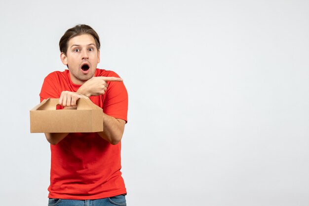 Вид сверху потрясенного и эмоционального молодого человека в красной блузке, держащего коробку, указывающего на что-то слева на белом фоне