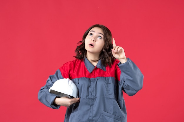 Free photo top view of shocked female builder in uniform and holding hard hat pointing up on isolated red background