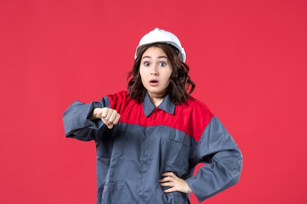 Foto gratuita vista dall'alto del costruttore femminile scioccato in uniforme con elmetto e che controlla il suo tempo su sfondo rosso isolato