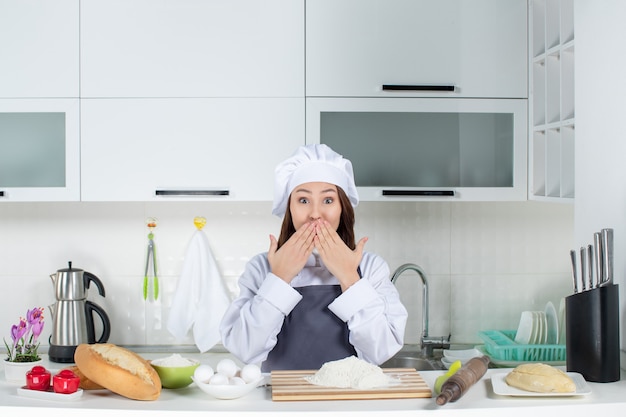 흰색 부엌에서 도마 빵 야채와 함께 테이블 뒤에 서 있는 제복을 입은 충격을 받은 여성 요리사의 상위 뷰