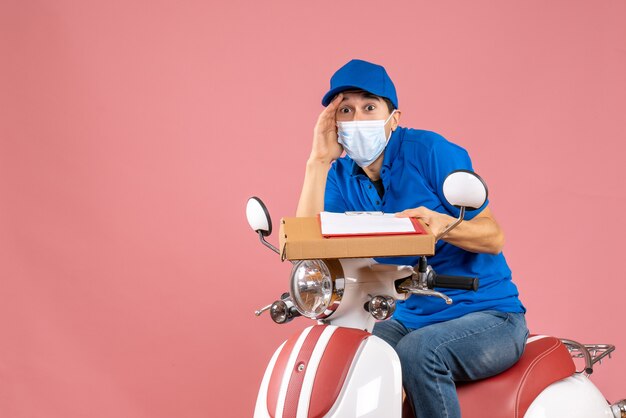 Vista dall'alto del fattorino maschio scioccato in maschera con cappello seduto su uno scooter che consegna ordini in possesso di documento su sfondo pesca