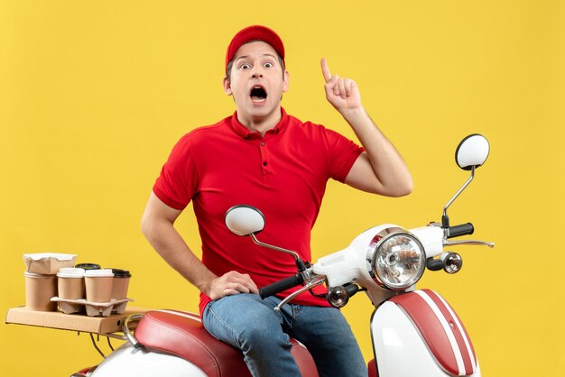 Top view of shocked young guy wearing red blouse and hat delivering orders pointing up on yellow background