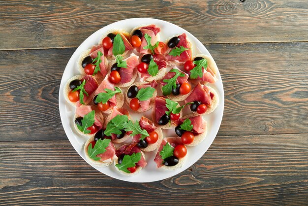 Top view shot of a big plate of ham canapes decorated with cherry tomatoes black olives and rucola leaves served on a wooden table copyspace restaurabt menu appetizers dish taste meal.