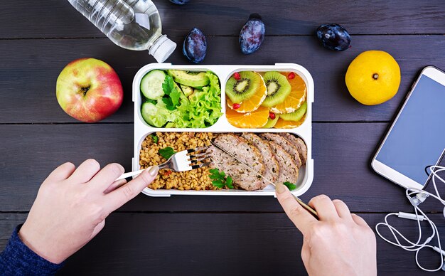 Взгляд сверху показывая руки есть здоровый обед с булгуром, мясом и свежими овощами и фруктами на деревянном столе. Фитнес и концепция здорового образа жизни. Коробка для ланча. Вид сверху