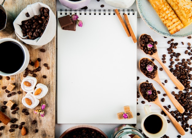 Top view of a sketchbook and coffee beans in wooden bowl and spoons and with lokum chocolate pieces and coffee beans scattered on rustic background