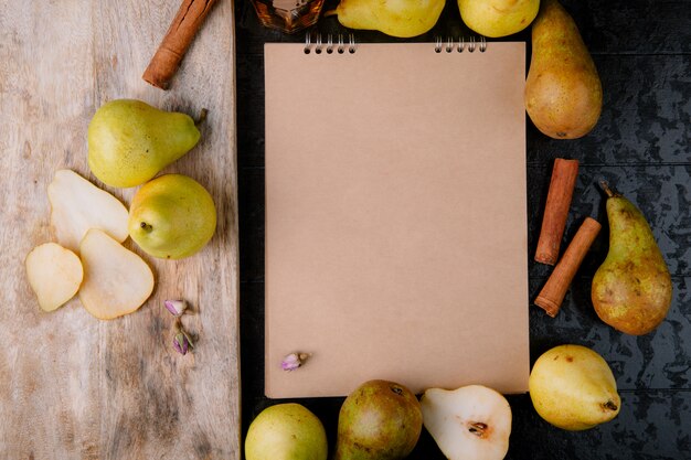 Top view of sketchbook made of craft paper framed with fresh ripe pears and a wooden chopping board with kitchen knife and slices of pears on black background