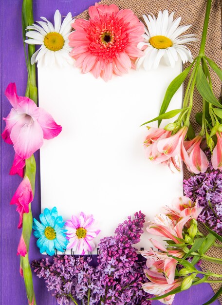 Top view of a sketchbook and pink and purple color flowers gerbera lilac alstroemeria and daisy flowers on sackcloth on purple wooden background