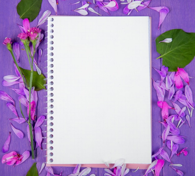 Top view of a sketchbook and purple flower petals scattered around on purple wooden background