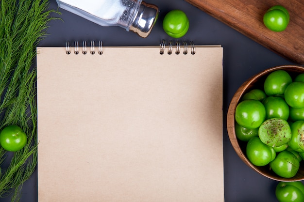 Top view of a sketchbook , salt shaker, fennel and sour green plums in a wooden bowl on black table