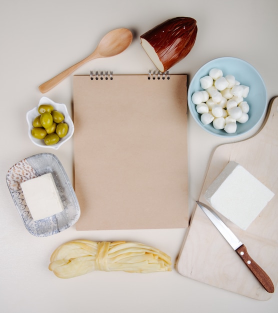 Free photo top view of a sketchbook and various types of cheese mini mozzarella cheese in a blue bowl , feta, smoked and string cheese with pickled olives on white table