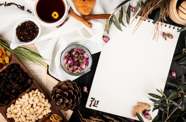 Top view of a sketchbook with tea rose buds in a glass jar, pine cones, mixed nuts and tree brunches with leaves and a cup of tea on black wood