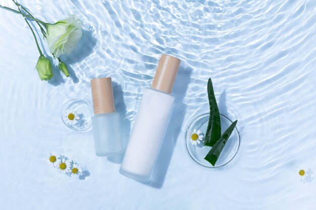 Top view of skincare bottles on a light blue water surface with aloe vera and daisy flowers