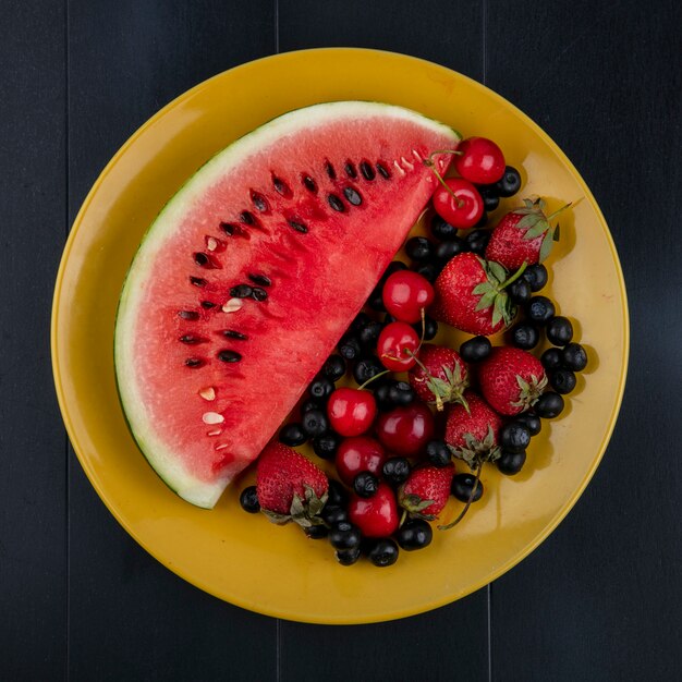 Top view slice of watermelon with cherries blueberries and strawberries on a yellow plate on a black background