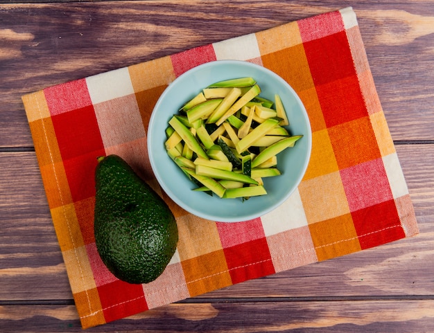 Top view of sliced avocado in bowl with whole one on cloth on wooden background