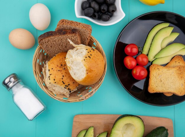 Top view of sliced avocados on a black plate with tomatoes and toasted bread with a bucket of breads with black olives on blue surface