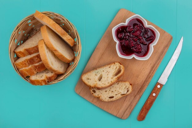 Top view of sliced baguette in basket and raspberry jam on cutting board with knife on blue background