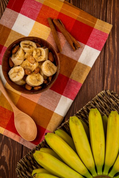 Вид сверху нарезанные бананы с миндалем в деревянной миске и кучу свежих бананов в плетеной корзине на деревенском