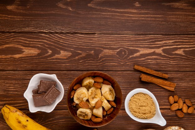 Top view of sliced bananas with almond in a wood bowl, dark chocolate and cinnamon sticks on wood rustic with copy space