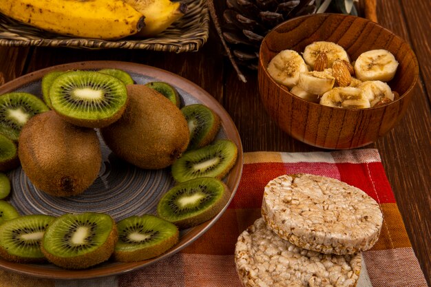 Top view of sliced bananas with almond in a wooden bowl, slices of kiwi fruit on a plate and rice crackers on rustic