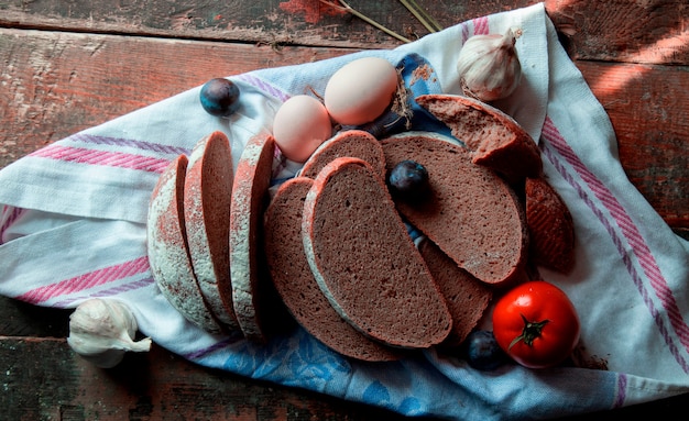 top view sliced black bread, eggs, plums, garlic gloves and tomatoes on a white tablecloth .