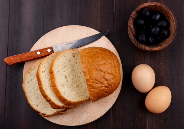 Вид сверху нарезанный хлеб и нож на разделочную доску с яйцами и миску черной оливы на деревянной поверхности