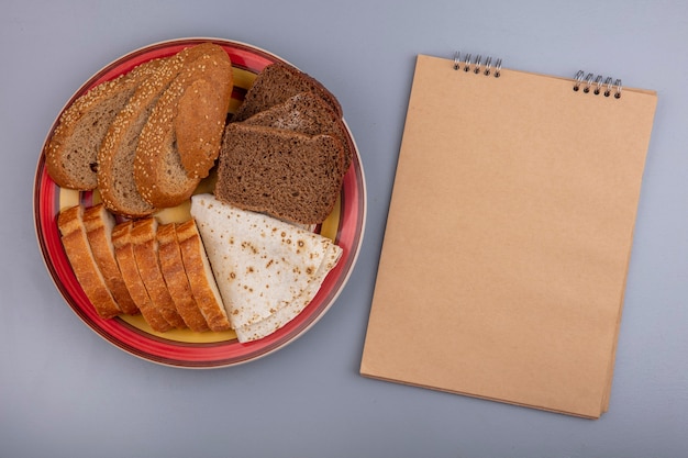 Вид сверху нарезанный хлеб в виде хрустящих лепешек из коричневых початков с семенами и ржаных в тарелке с блокнотом на сером фоне с копией пространства