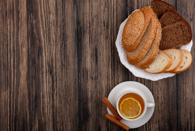 Top view of sliced breads as seeded brown cob rye and white ones in plate and cup of hot toddy with cinnamon on saucer on wooden background with copy space