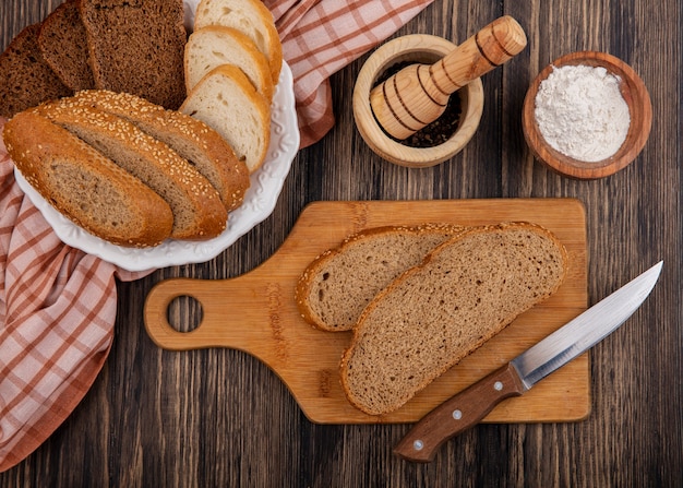 Top view of sliced breads as seeded brown cob rye and white ones in plate on plaid cloth and on cutting board with knife black pepper and flour on wooden background