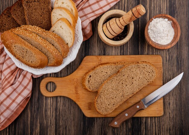 Top view of sliced breads as seeded brown cob rye and white ones in plate on plaid cloth and on cutting board with knife black pepper and flour on wooden background