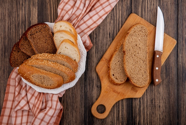 Top view of sliced breads as seeded brown cob rye and white ones in plate on plaid cloth and on cutting board with knife on wooden background