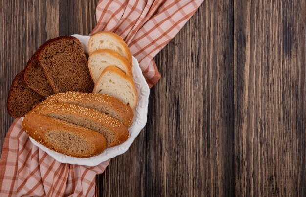 Top view of sliced breads as seeded brown cob rye and white ones in plate on plaid cloth on wooden background with copy space