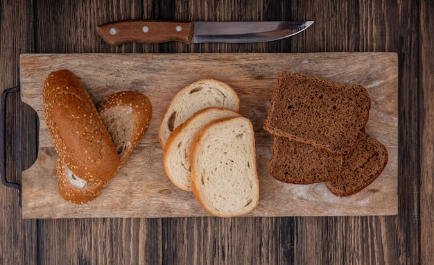 Vista dall'alto di fette di pane come pannocchia marrone seminato bianco e quelli di segale sul tagliere e coltello su sfondo di legno