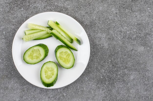 Top view of sliced and chopped cucumber slices on white plate over grey table.